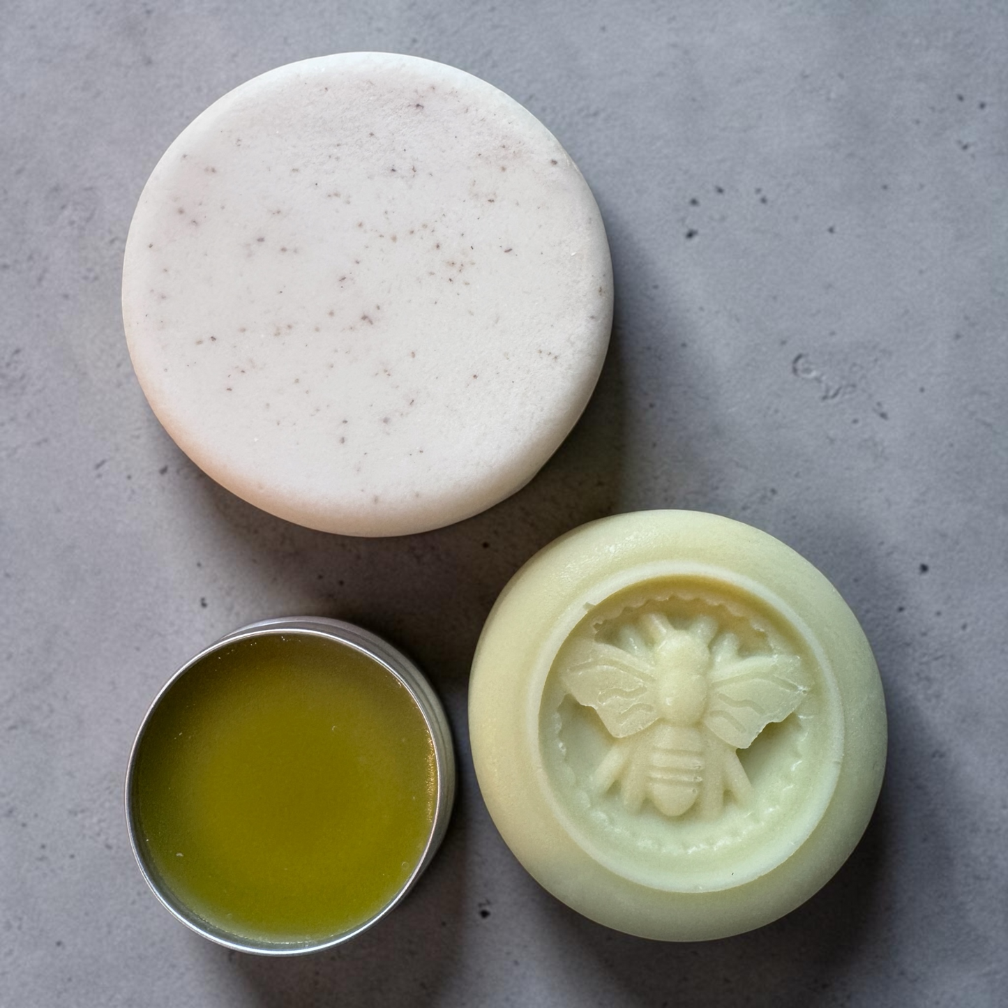 A round white bar of soap with fine dark specks, shown next to a small metal tin filled with yellow-green balm and a pale yellow beeswax mold featuring an embossed bee design, all arranged on a gray textured surface.