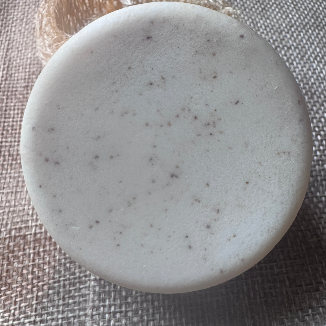 A round white bar of soap with small dark speckles, photographed from above on a textured gray woven surface.